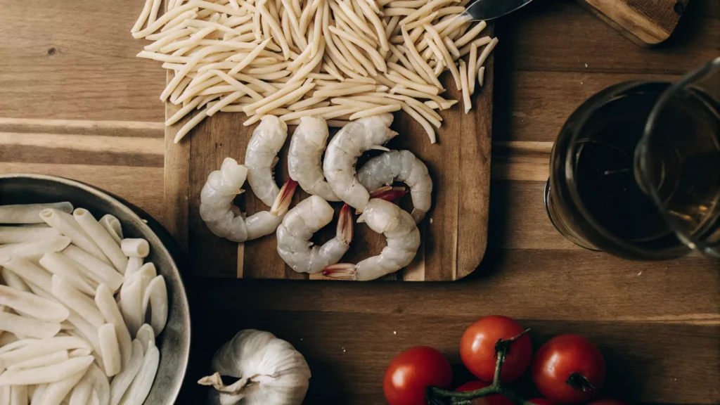 A flat-lay image of ingredients for Strozzapreti with Jumbo Shrimp, featuring strozzapreti pasta, peeled jumbo shrimp, minced garlic, olive oil, halved cherry tomatoes, fresh basil, grated Parmesan cheese, red pepper flakes, and salt and pepper on a rustic wooden surface.